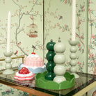 Een decoratieve tafel met Addison Ross Bobbin kandelaars met witte kaarsen, een groene Addison Ross Bobbin Zout-en Pepermolen Racing Groen 24 cm, twee desserts, alles tegen vogel- en takkenbehang met een vogelkooi.