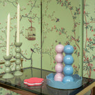 Een pastelkleurige tafelschikking met twee hoge witte kaarsen in lichtgroene houders, een roze bloemenonderzetter en de Addison Ross Bobbin Zout-en Pepermolen Roze 24 cm met keramisch maalwerk op een blauw dienblad tegen groen vogel- en takbehang.