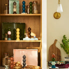 Op houten planken staan potten, schalen en kleurrijk gestapelde Addison Ross Bobbin zout- en pepermolen in Racing Groen (24 cm). Onder de tafel staan brood, extra molens, snijplanken en een plant. Addison Ross borden zijn zichtbaar op de achtergrond.