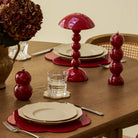 Een houten gedekte eettafel met crèmekleurige borden op rode placemats, heldere glazen, een Addison Ross Bobbin Lamp LED Oplaadbaar Kersenrood, bijpassend rood peper- en zoutstel en een vaas met hortensia's.