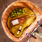 Een rieten picknickmand met een fles rosé, groene druiven, een Addison Ross Bowtie Kurkentrekker Amie kurkentrekker en een kurk ligt op een gele doek bovenop een houten oppervlak.