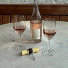 Een fles rosé met een minimalistisch etiket staat op een marmeren tafel met twee glazen. Vlakbij liggen een open kurk, een gesloten kurk en de Addison Ross Strik Kurkentrekker Amie. Daarachter liggen houten lambriseringen en een witte muur.