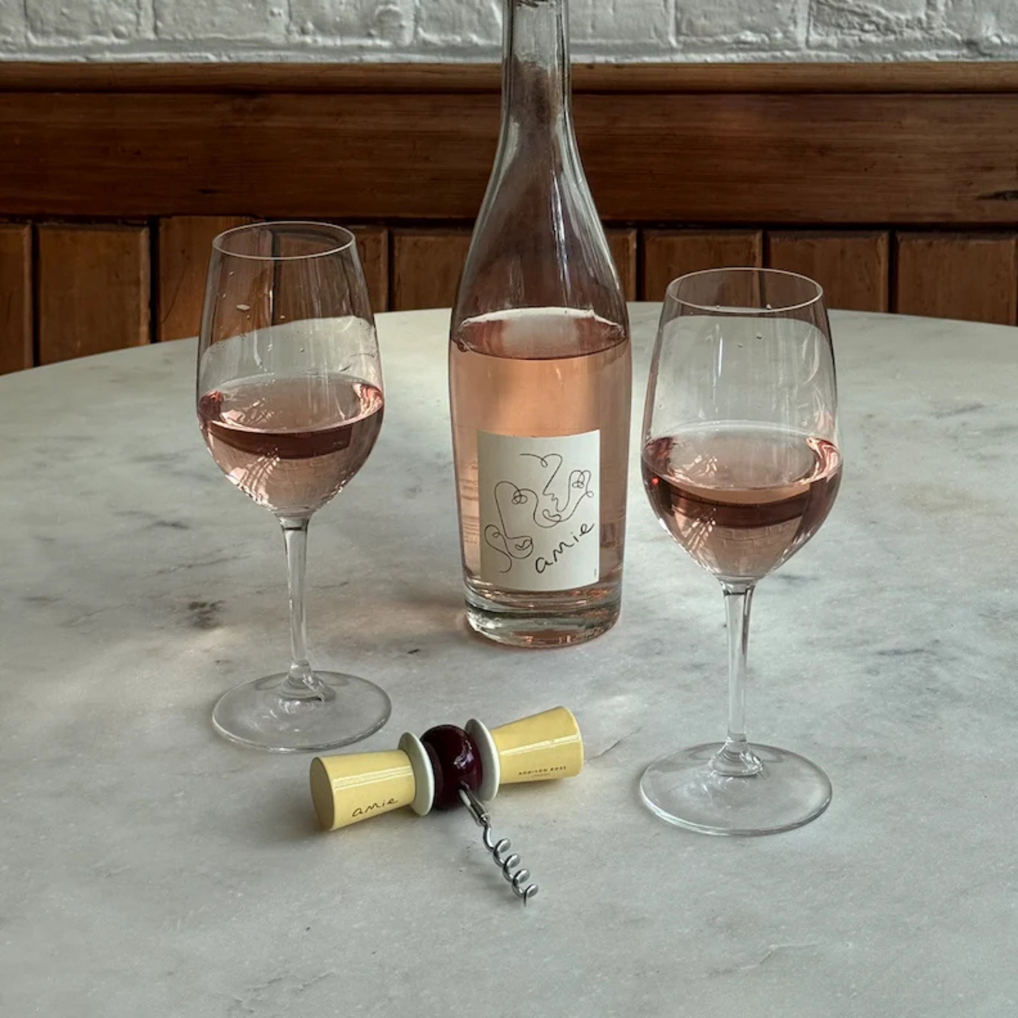 Een fles rosé met een minimalistisch etiket staat op een marmeren tafel met twee glazen. Vlakbij liggen een open kurk, een gesloten kurk en de Addison Ross Strik Kurkentrekker Amie. Daarachter liggen houten lambriseringen en een witte muur.