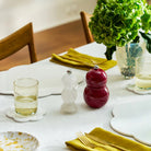 Een gedekte eettafel met gele servetten, borden, glazen water, een Addison Ross Chubby Zout-en Pepermolen Kersenrood 12 cm, en een vaas met groene hortensia's, met houten stoelen op de achtergrond.