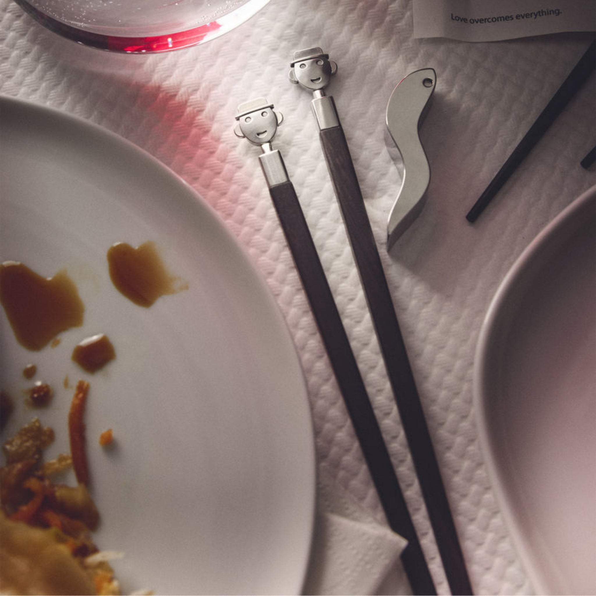 Twee Alessi Alessandro eetstokjes + Houder van Alessi, met cartoonachtige gezichten geïnspireerd door Mendini, rusten op een wit oppervlak met textuur naast tafelaccessoires: een zilveren flesopener, een bord met eten en saus, een rood drankje in een glas en een zichtbare kaart.