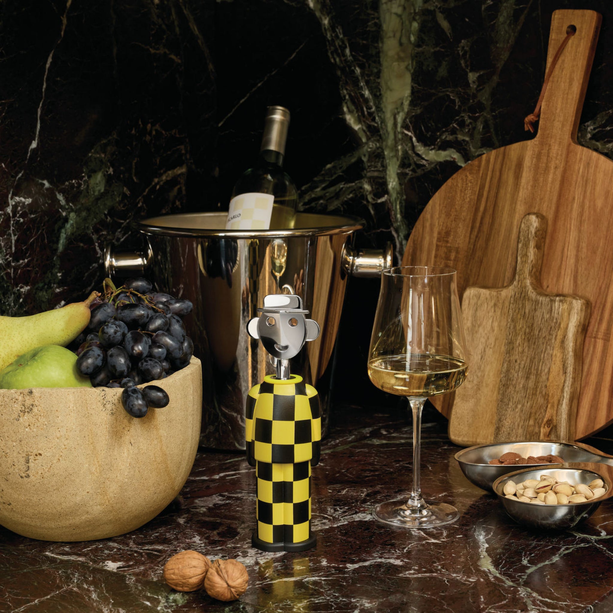 Een Alessi Alessandro Kurkentrekker Giallo e Nero, een opvallend Italiaans designicoon, staat op een marmeren toonbank naast fruit, een wijnglas, witte wijn in een ijsemmer, houten planken en schaaltjes noten.
