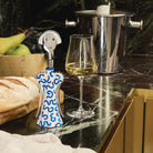 Een Alessi Anna G Kurkentrekker Delft kurkentrekker, een glas witte wijn, brood, fruit en een wijnemmer met een fles staan opgesteld op een donker marmeren aanrecht.