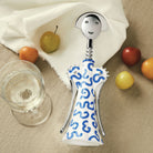 De Alessi Anna G Kurkentrekker Delft van Alessi, met een lachend gezichtje en Delftsblauw ontwerp, rust op een beige tafelkleed naast een glas witte wijn, een wit servet en verschillende kleurrijke vruchten.