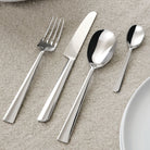 Vier glanzend zilveren bestekken uit de Alessi Asta Bestek 24-delig set van Alessi-een vork, mes, eetlepel en theelepel-zijn netjes geplaatst op een beige tafelkleed met textuur naast een wit bord.