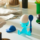 Een zachtgekookt eitje zit in het speelse Alessi Cico Eierdopje Blauw van Alessi, in de vorm van een octopus en met een bijpassende lepel in de hand. De blauwe dop rust vlakbij op een zonovergoten aanrecht.