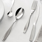 Een Alessi Duna Bestek 24-delig servies met Italiaans design staat op een wit tafelkleed naast een bord en servet.