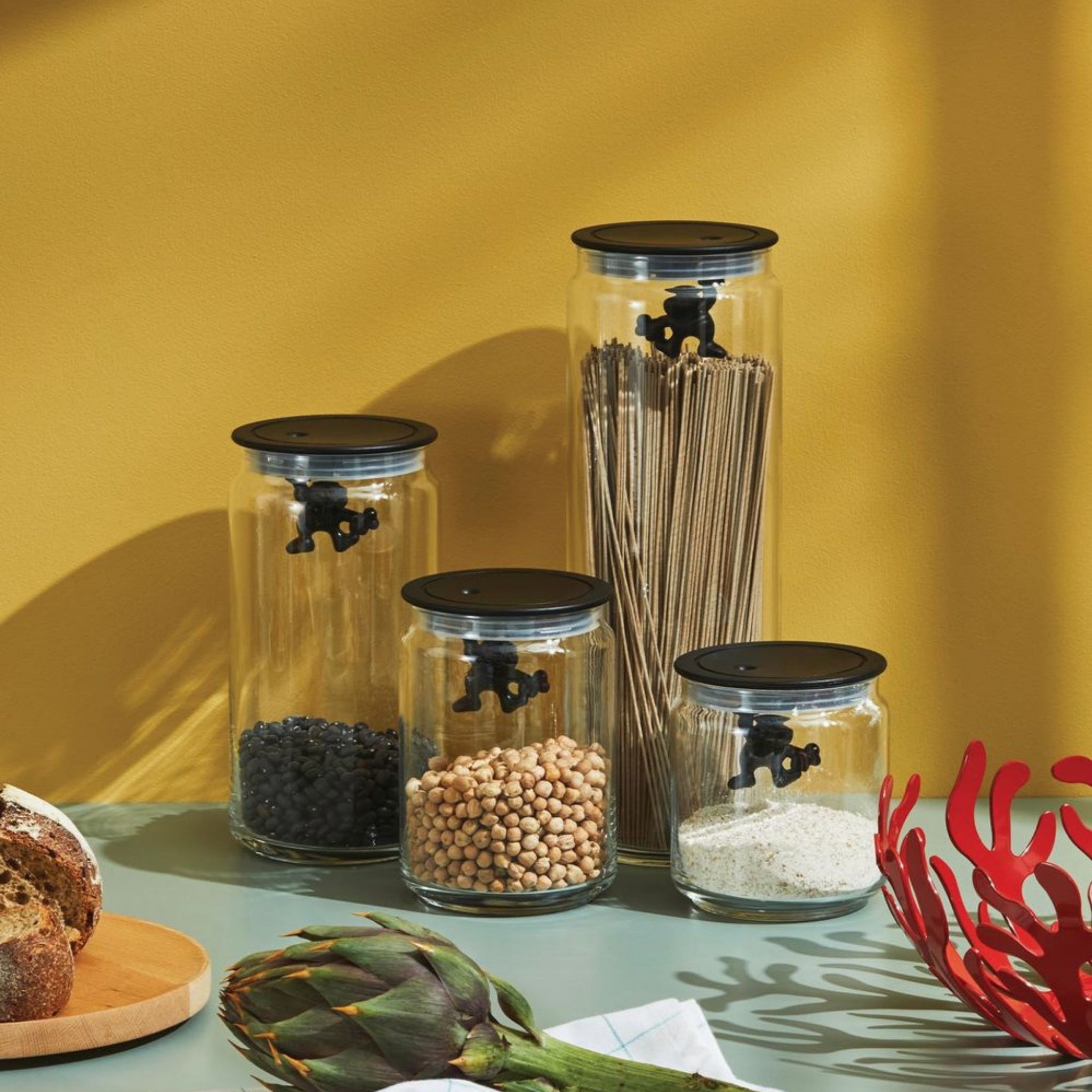 Vier glazen potten met zwarte deksels, waaronder de Alessi Gianni Voorraadpot Zwart 90 cl, houden zwarte bonen, kikkererwten, rijst en spaghetti op een aanrecht tegen een gele muur voor een stijlvol keukenaccent.