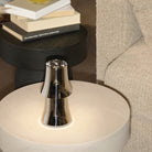 De Alessi Halesia Tafellamp LED Oplaadbaar staat op een ronde, lichte bijzettafel naast een beige bank met textuur, terwijl gestapelde boeken op een zwart tafeltje op de achtergrond rusten en een gezellige en eigentijdse sfeer creëren.