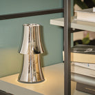 De Alessi Halesia Tafellamp LED Oplaadbaar, met zijn strakke zilveren afwerking, staat op een witte plank naast boeken. Deze moderne lamp van Alessi werpt een zachte, warme gloed en brengt elegantie in elke moderne woonkamer of kantoor.