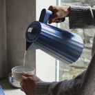 Een persoon giet heet water uit de Alessi Plissé Elektrische Waterkoker Blauw 1,7 l in een witte mok bij een raam, met de hand en een deel van een grijze trui zichtbaar.