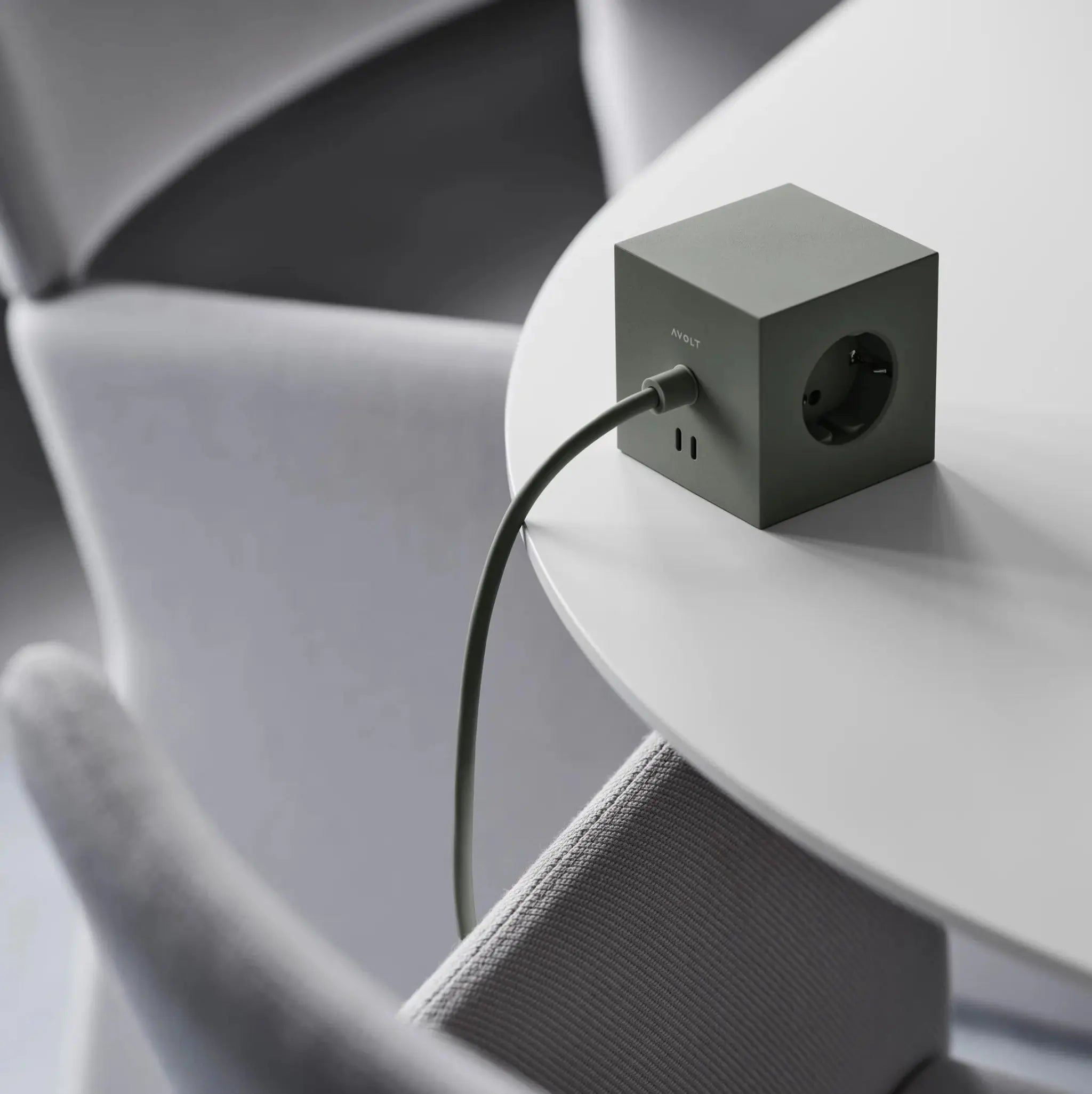 Een Avolt Square 1 USB-C Stekkerdoos Eik Groen 1,8 m van Avolt ligt op een witte ronde tafel, met het snoer aangesloten. Lichtgrijze stoelen omringen de opstelling en creëren een moderne en minimalistische sfeer.