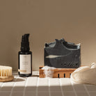 Een flesje huidverzorgingsserum, een Azur Black Beauty Natuurlijke Gezicht Zeep Bar op een houten houder met schuim, een gezichtsborstel en een zachte doek zijn gerangschikt op tegels onder warm licht voor een Azur plasticvrije routine.