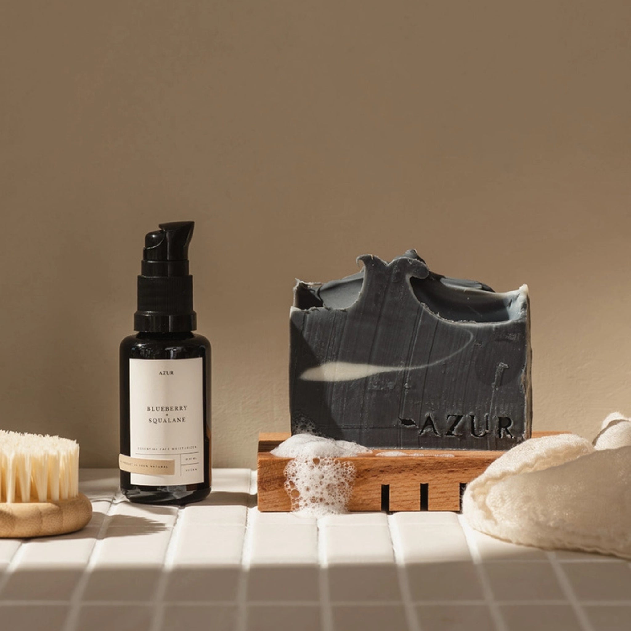 Een flesje huidverzorgingsserum, een Azur Black Beauty Natuurlijke Gezicht Zeep Bar op een houten houder met schuim, een gezichtsborstel en een zachte doek zijn gerangschikt op tegels onder warm licht voor een Azur plasticvrije routine.