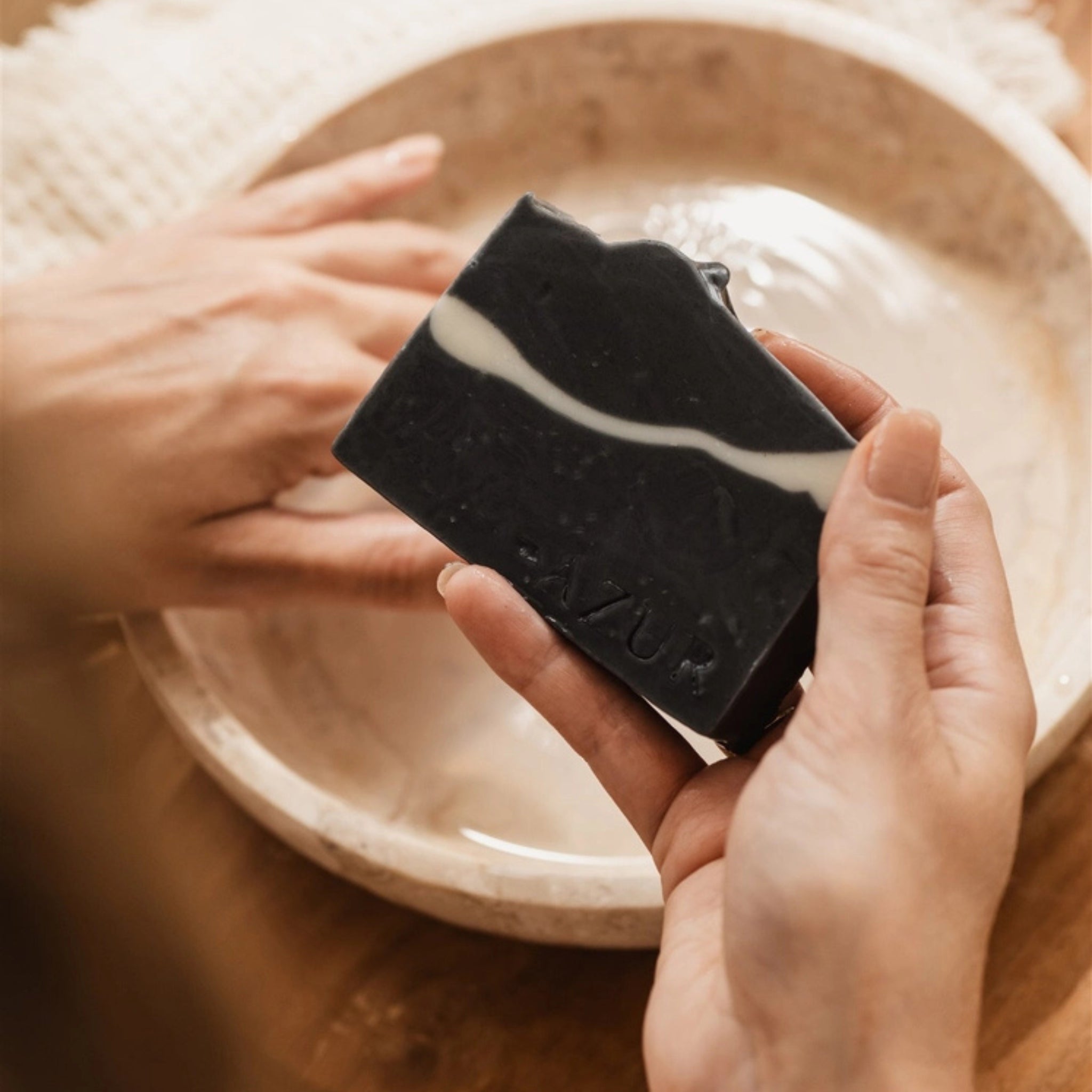 Een persoon houdt een Azur Black Beauty Natuurlijke Gezichtszeepstaaf boven een ondiepe kom water, terwijl een andere hand op de rand van de kom rust, ter voorbereiding op het wassen met deze gezichtsreiniger van Azur.