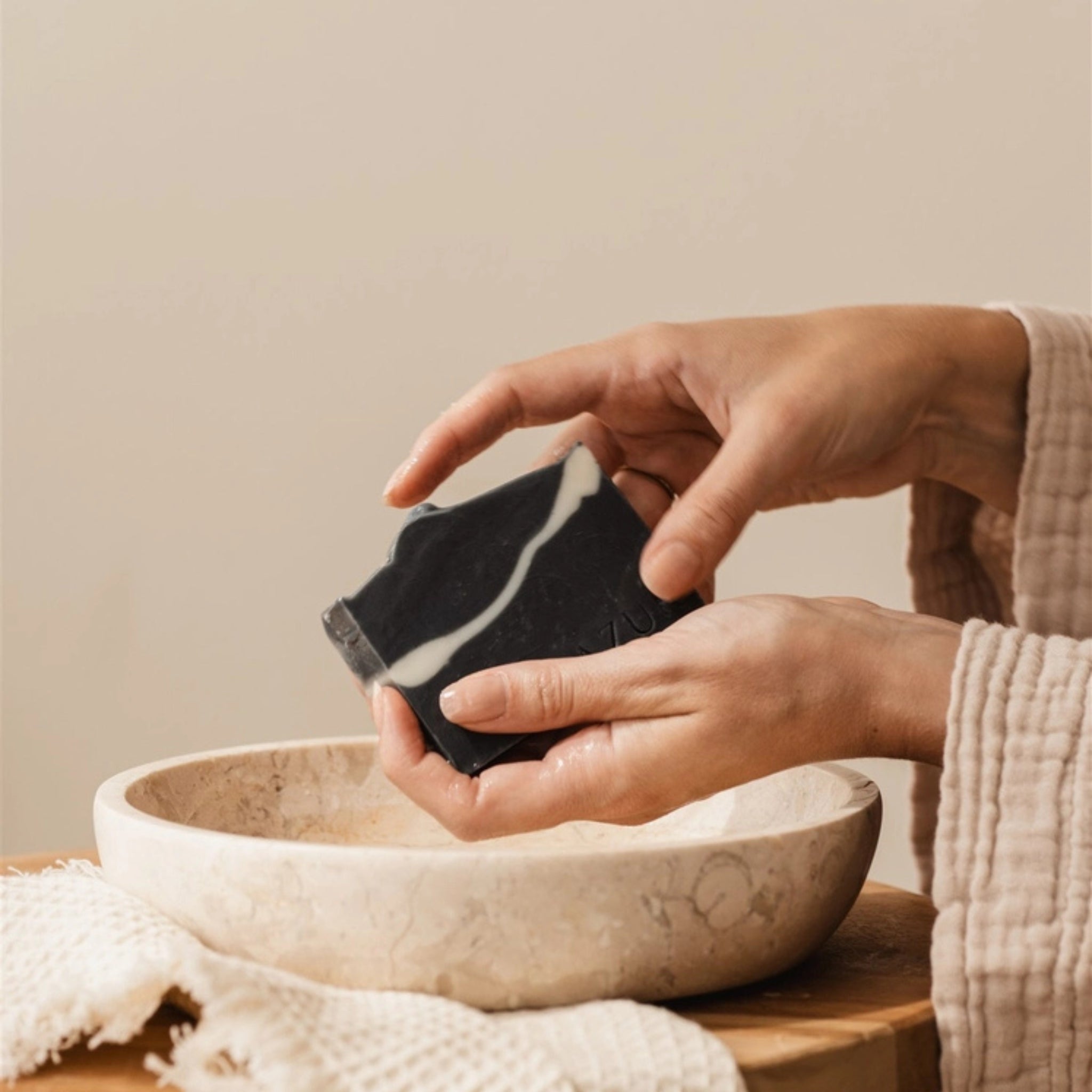 Een persoon in een beige gewaad houdt een Azur Black Beauty Natuurlijke Gezicht Zeep Bar van Azur boven een ronde, lichtgekleurde stenen kom, met een gestructureerde doek in de buurt op een houten oppervlak.
