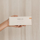 Een hand houdt een rechthoekige beige doos vast met de tekst "Azur Sample Set Natuurlijke zepen 4 st." van Azur, met natuurlijke zepen tegen een neutrale, lichtgekleurde stoffen achtergrond.