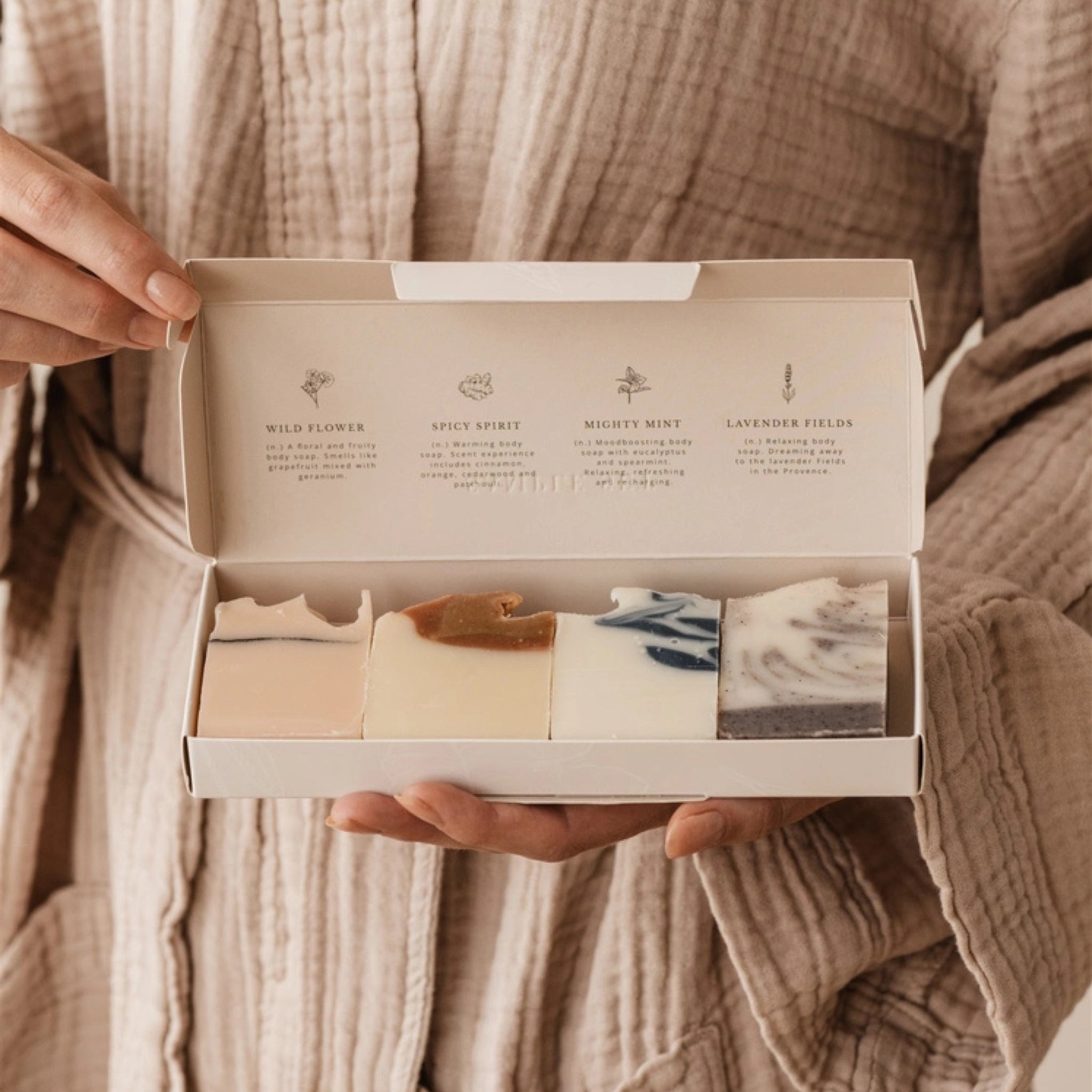 Een persoon in een beige badjas houdt een open Azur Sample Set Natuurlijke zepen 4 st. doos vast, waarop vier kleurrijke, plasticvrije bodybars van Azur staan met de namen Wild Flower, Spicy Spirit, Mighty Mint en Lavender Fields.