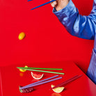 Een persoon in een blauwe satijnen hoes gebruikt Byon Yaki Chopsticks 4 st. van Byon boven een rode tafel met kleurrijke eetstokjes, speelgoedfruit en een drijvende citroen - alles tegen een levendige rode achtergrond, waardoor het een ideaal uniek cadeau is.