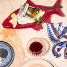 Een Byon Kai Serveerschaal Rood van Byon in de vorm van een vis toont een open blikje sardines met citroenpartjes en kruiden. Een vork rust op het blikje. Vlakbij: een glas rode wijn, zwart bord met kruimels, servet en beige bord op marmer.