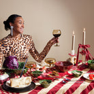 Een vrouw in een topje met luipaardprint lacht terwijl ze een wijnglas vasthoudt aan een tafel met kaarsen, kaas en groenten geserveerd op de Byon Kai Serveerschaal Rood van Byon, gedekt op een rood en beige geruit tafelkleed.