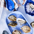 Het Byon Oester Schaaltje Blauw van Byon houdt oesters op ijs bovenop een wit oppervlak, met meer oesters en ijs in andere blauwe schaaltjes in de buurt en een gedessineerd doek op de achtergrond.