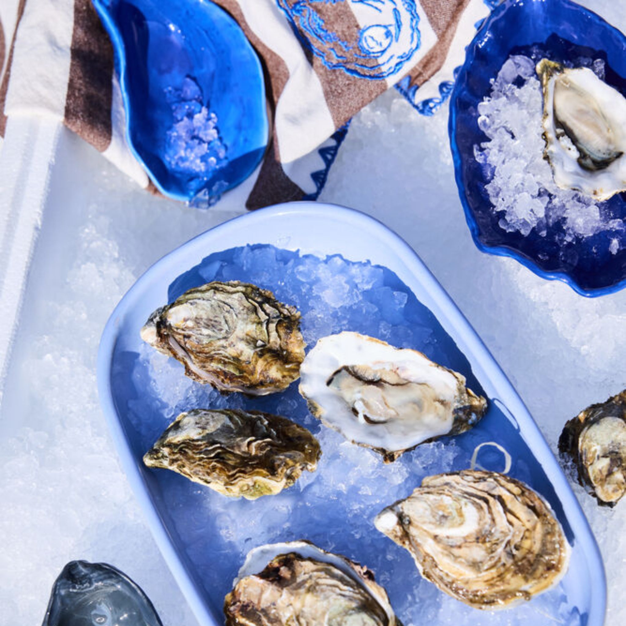 Het Byon Oester Schaaltje Blauw van Byon houdt oesters op ijs bovenop een wit oppervlak, met meer oesters en ijs in andere blauwe schaaltjes in de buurt en een gedessineerd doek op de achtergrond.