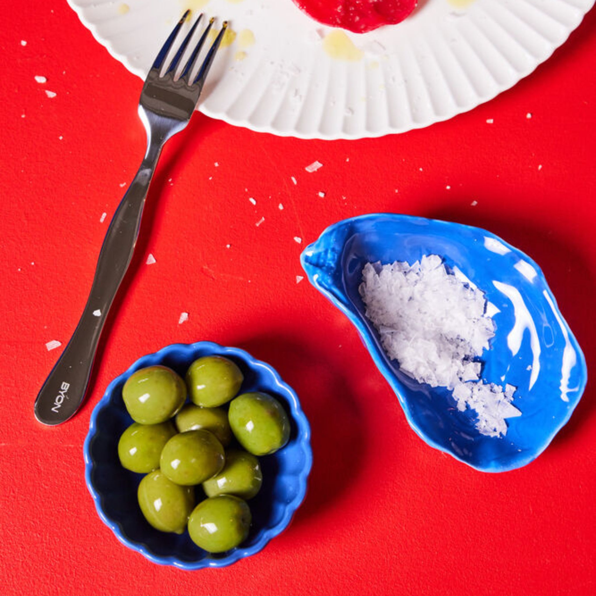 Een Byon Oester Schaaltje Blauw met groene olijven en een ander met vlokkig zout rusten op een rode tafel naast een vork en een wit bord met etensresten.