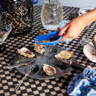 Een hand giet saus over oesters geserveerd in het Byon Oester Schaaltje Blauw op een donker bord met drie vorken. Het geruite tafelkleed is gedekt met drankjes, glaswerk en een blauw servet.