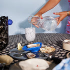 Een persoon schenkt water uit een heldere kan in een wit gemarmerd kopje op een geruite tafel met oesters, citroenpartjes, verschillende schaaltjes en het Byon Oester Schaaltje Wit van Byon, wat bijdraagt aan de ongedwongen zomerse sfeer.