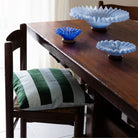 Een houten eettafel met drie glazen schalen met blauwe ruches staat naast een houten stoel met de Byon Pathi Kussen Groen/Blauw 45/45, terwijl het licht door een gordijn op de achtergrond valt.