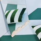 Een geschulpte gouden spiegel weerspiegelt een Byon Pathi Kussen Groen/Blauw 45/45 kussen, gemaakt van zacht katoenen velours, tegen een groen-wit geblokte vloer. Een ander bijpassend Byon kussen ligt vlakbij.