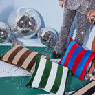 Een feestelijk tafereel met drie Byon Pathi Kussen Groen/Blauw 45/45 in trendy strepen, zilveren discoballen en metallic confetti. Twee mensen in paillettenoutfits staan vlakbij en laten alleen hun glitterbenen zien; een van hen houdt een glas wijn vast.