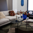 Een moderne woonkamer is voorzien van een lichte bank met structuurkussens, een zwarte salontafel met boeken, de Byon Ruby Kandelaar Groen/Blauw van Byon, blauwe vazen met rode bloemen, abstracte decoratie en een vloerkleed met patroon.