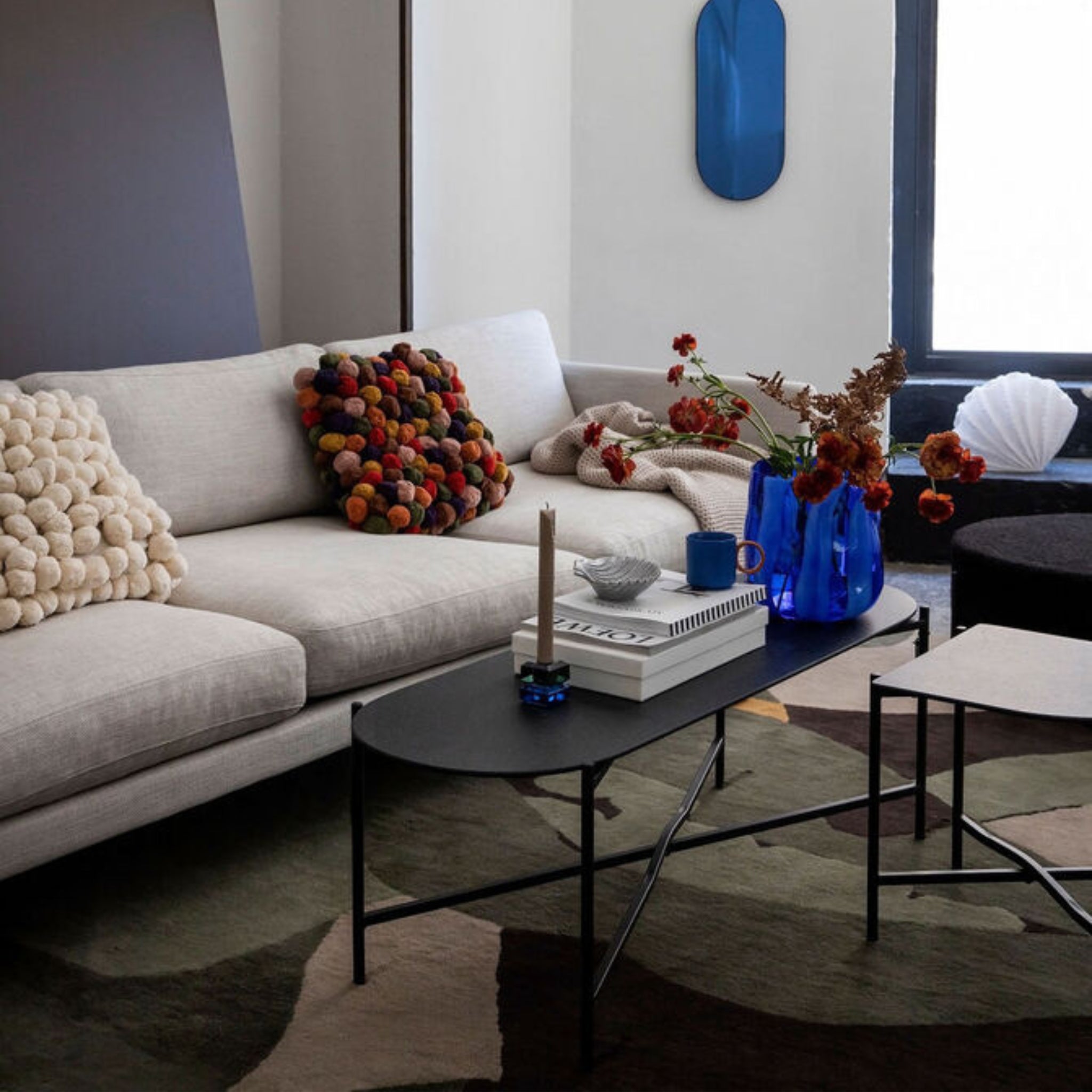 Een moderne woonkamer is voorzien van een lichte bank met structuurkussens, een zwarte salontafel met boeken, de Byon Ruby Kandelaar Groen/Blauw van Byon, blauwe vazen met rode bloemen, abstracte decoratie en een vloerkleed met patroon.