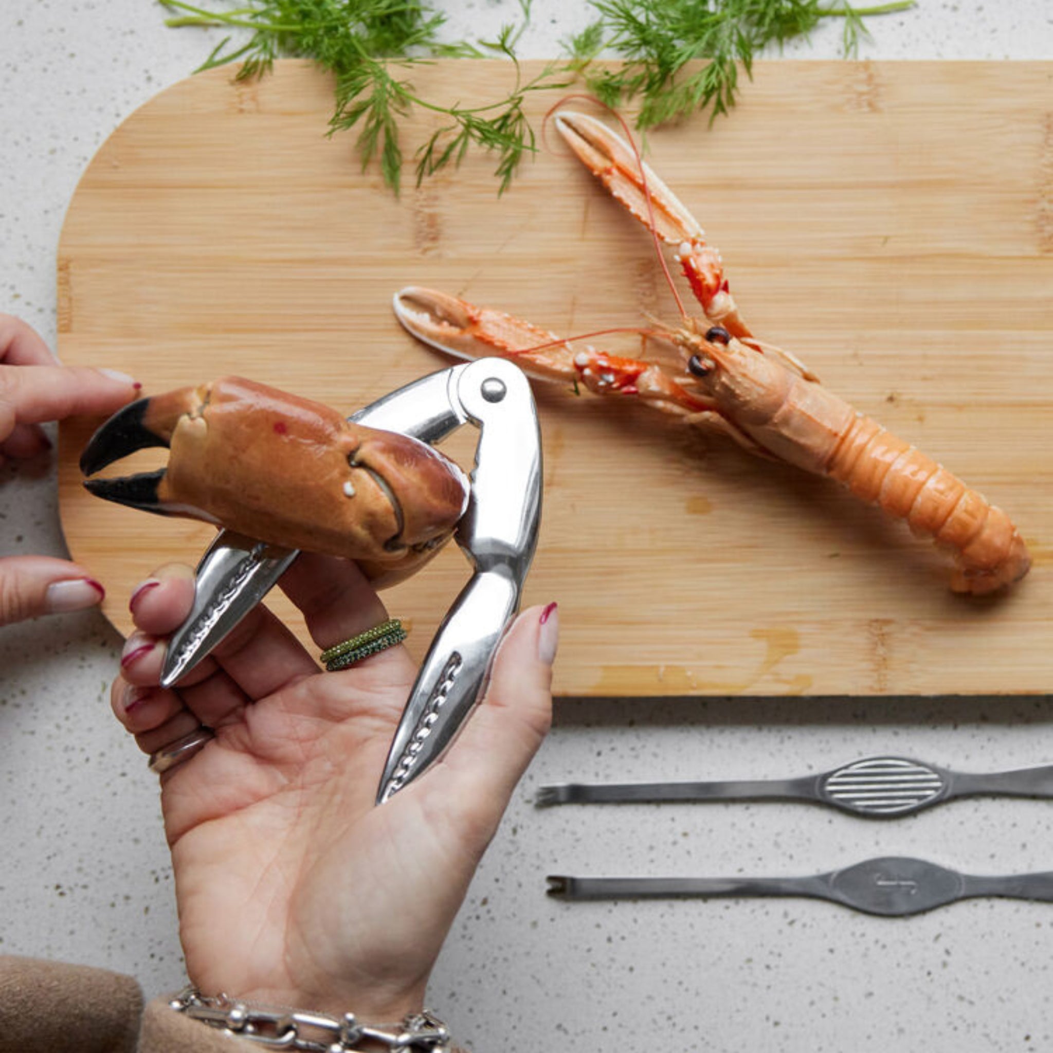 Een persoon gebruikt de Byon Sivan Schelpdier/Notenkraker van Byon, gemaakt van roestvrij staal, om een bruine krabklauw te openen op een houten snijplank, met een langoustine, dille en zeevruchtenplukken op de achtergrond.