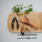 Een Byon Sivan Schelpdier/Notenkraker van Byon met twee zeevruchtenprikkers rust op een houten plank naast verse dille, een kreeftenklauw en een krabklauw op een wit gespikkeld aanrecht.