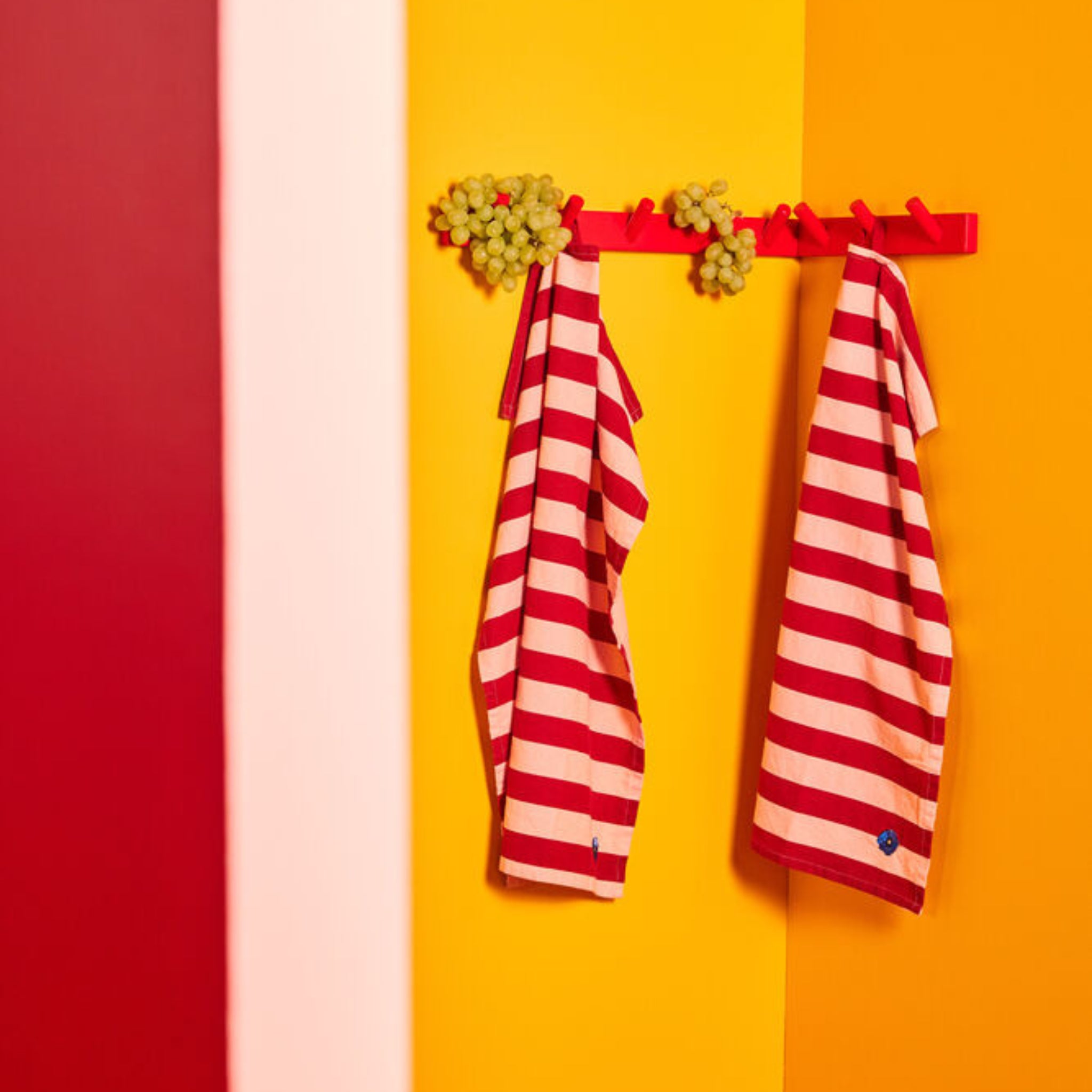 Een rode kapstok met groene druiven en twee Byon Theedoek Leya Rood 2 st. hangen aan een muur gestreept in rood, roze, geel en oranje. Elke Byon keukendoek is gemaakt van biologisch katoen.
