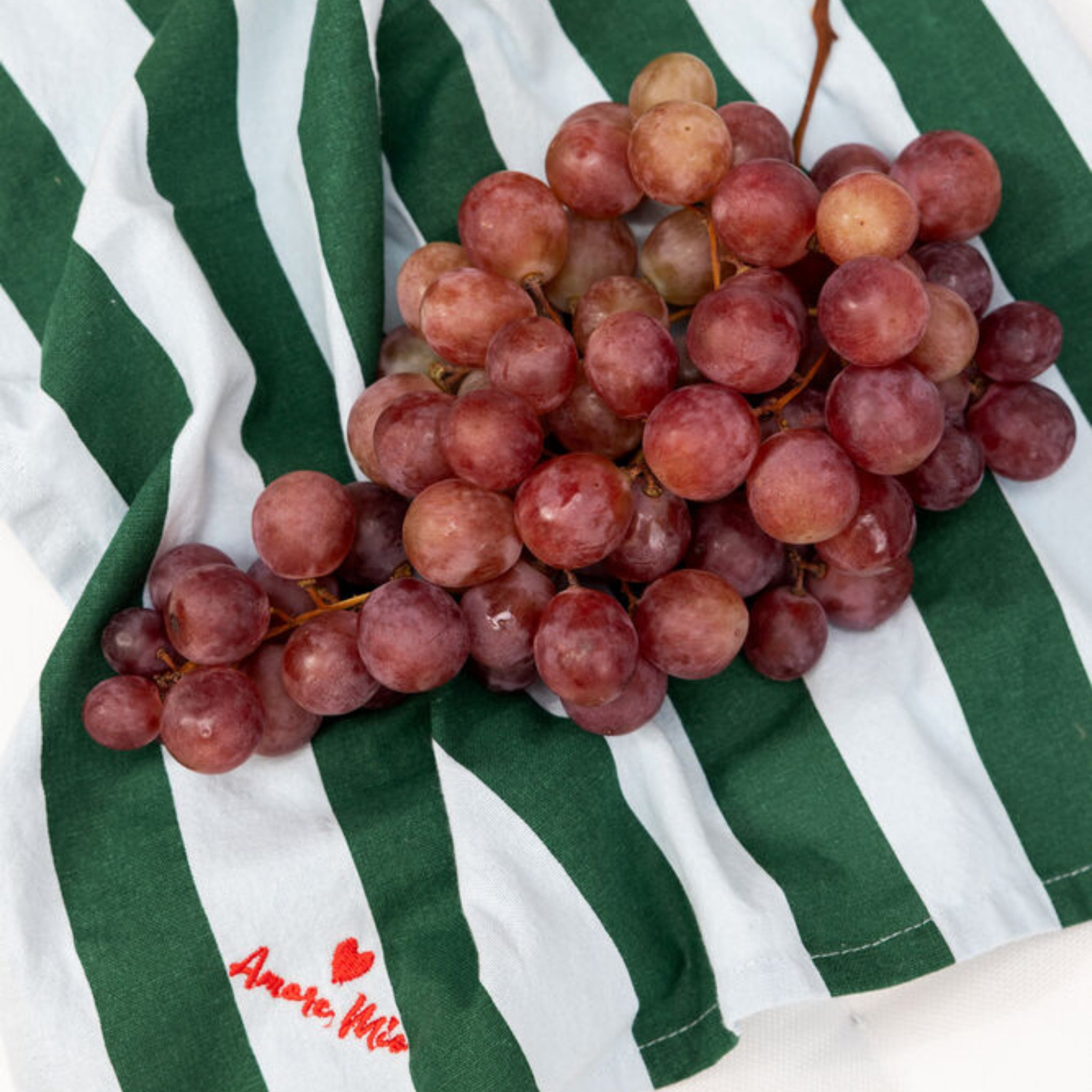 Een tros rode druiven rust op een Byon Theedoek Love Groen/Blauw 2 st. van Byon, gemaakt van biologisch katoen met een groen en wit streepdessin, een klein rood hartje en 'Amore, Milan' borduursel - een charmant keukencadeau.
