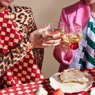 Twee mensen toosten met witte wijn op een Byon Theedoek Love Rood van Byon, omringd door brood en kleine kalebassen. De een draagt luipaardprint en ruitjes, de ander roze en groene strepen, die kleur toevoegen aan het vrolijke tafereel.