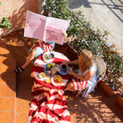 Twee mensen zitten aan een kleine buitentafel met ontbijtartikelen en een Byon Theedoek Leya Rood 2 st. gemaakt van biologisch katoen. Eén van hen houdt een groot roze bord vast dat hun gezicht gedeeltelijk bedekt terwijl het zonlicht schaduwen werpt op het terras. Merk: Byon.