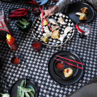 Een hand met rode Byon Yaki Chopsticks 4 st. reikt naar voedsel op een geruite schotel, omringd door borden, saus, rode pepers en sriracha-een onderscheidend cadeau van Byon voor elke liefhebber van de bamboekeuken.