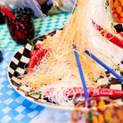 Een bord dunne, knapperige noedels met rode chilipepers wordt geserveerd met Byon Yaki Chopsticks 4 st. van Byon op een blauw-wit geblokt tafelkleed tussen kleurrijke gerechten - een perfect cadeau voor elke liefhebber van bamboe.