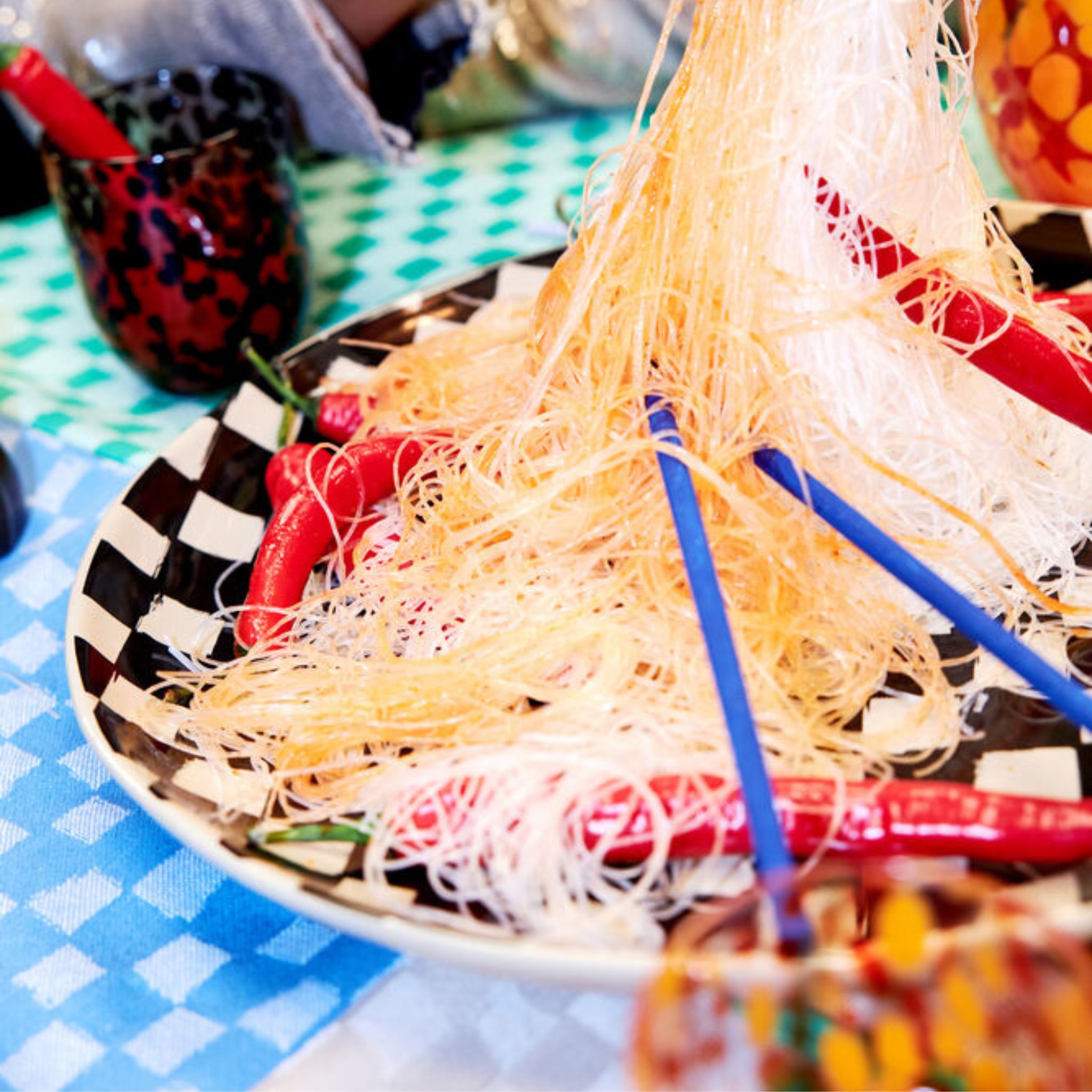 Een bord dunne, knapperige noedels met rode chilipepers wordt geserveerd met Byon Yaki Chopsticks 4 st. van Byon op een blauw-wit geblokt tafelkleed tussen kleurrijke gerechten - een perfect cadeau voor elke liefhebber van bamboe.
