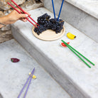 Een hand plukt druiven van een bord op een marmeren trap met Byon Yaki Chopsticks 4 st. van Byon, terwijl kleurrijke bamboe eetstokjes en kleine fruitvormige cadeau-artikelen in de buurt verspreid liggen.