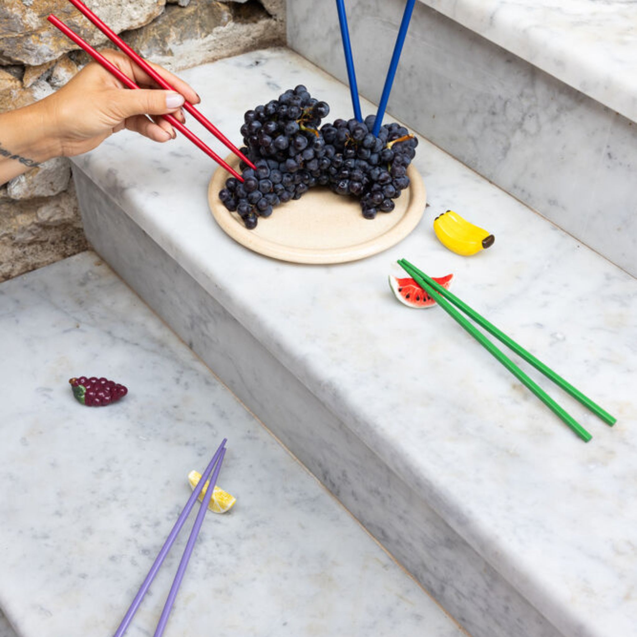 Een hand plukt druiven van een bord op een marmeren trap met Byon Yaki Chopsticks 4 st. van Byon, terwijl kleurrijke bamboe eetstokjes en kleine fruitvormige cadeau-artikelen in de buurt verspreid liggen.